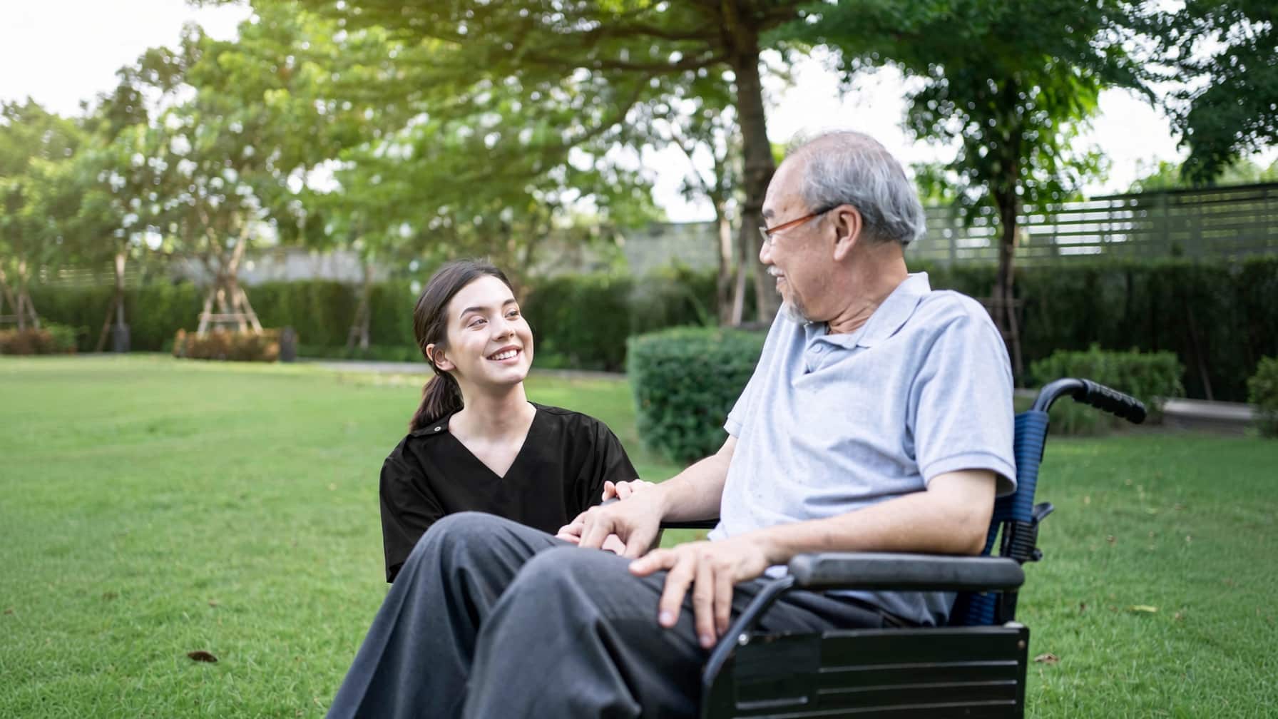 Caregiver in black scrubs sits outdoors with an elderly man in a wheelchair, smiling and holding his hand in a sunny park.