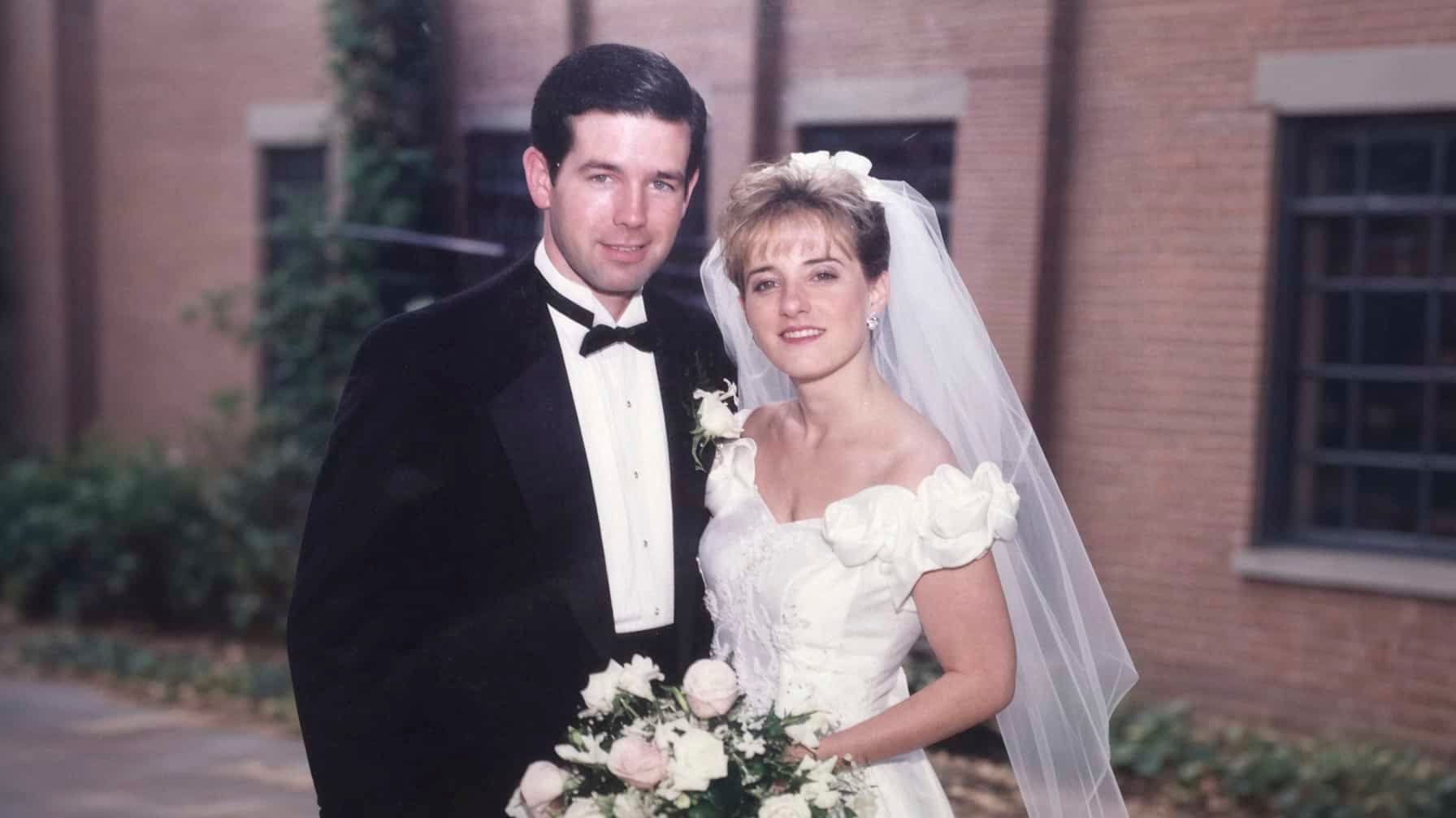 Dan and April Poch on their wedding day in the 1990s, he in a black tuxedo and she in a white gown and veil holding a bouquet of roses.